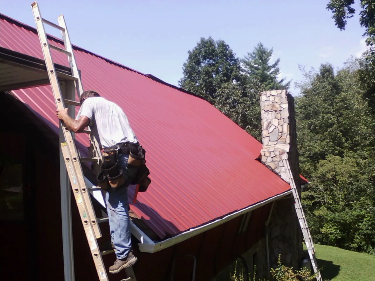 Roofer on a ladder beside a red metal roof and stone chimney — Soffit & Fascia Repair by Roofing Brothers in Branford